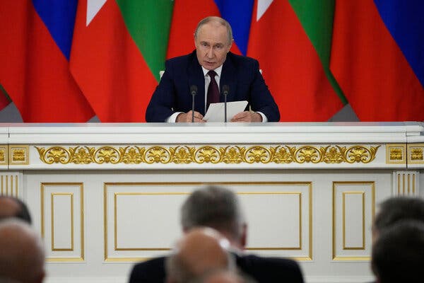 Tổng thống Vladimir V. Putin của Nga ngồi sau một chiếc bàn trắng được trang trí mạ vàng, trước một hàng cờ, đang phát biểu vào micro. Mọi người ngồi ở phía trước, đối diện với ông.
